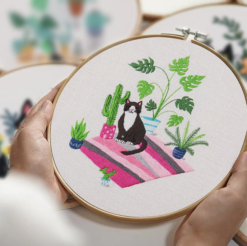 May include: Embroidered hoop art featuring a black and white cat sitting on a pink and gray striped rug surrounded by potted plants, including a cactus and a monstera plant.