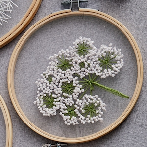 May include: A white and green embroidered flower design in a wooden embroidery hoop. The flower is a delicate white with green stems and leaves.