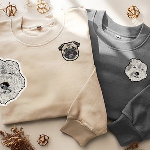 Op de afbeelding: Twee sweatshirts met geborduurde honden gezichten. Een sweatshirt is beige met een wit honden gezicht en de andere is grijs met een mops gezicht.