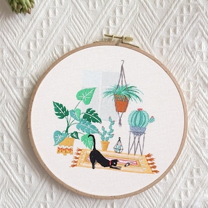 May include: Embroidered hoop art featuring a black cat, potted plants, and a cactus on a rug. The design is stitched on a white fabric background.
