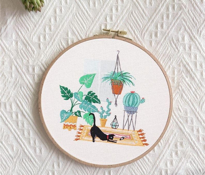 May include: Embroidered hoop art featuring a black cat, potted plants, and a cactus on a rug. The design is stitched on a white fabric background.