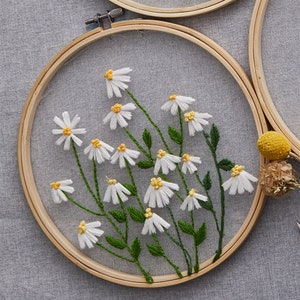 May include: A close-up of a white daisy embroidery hoop art piece. The hoop is made of wood and has a white mesh fabric stretched over it. The daisies are embroidered in white thread with yellow centers. The embroidery is surrounded by green leaves.
