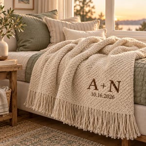 Könnte beinhalten: Eine cremefarbene, grob gestrickte Wohndecke mit langen Fransen. Die Decke ist mit dunkelbraunem Text bestickt, der A + N und das Datum 10.16.2026 zeigt. Der strukturierte Stoff liegt auf einem Bett mit verschiedenen Kissen im Hintergrund.