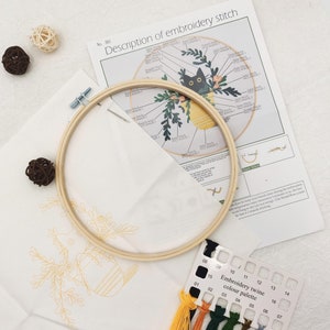 May include: A white embroidery hoop with a black cat and floral design on the fabric. The hoop is surrounded by embroidery floss in various colors, including yellow, orange, green, and blue. The floss is labeled with numbers 01-16. The image also includes a sheet of instructions with the text "Description of embroidery stitch" and "Embroidery twine color palette".