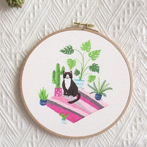 May include: Embroidered hoop art featuring a black and white cat sitting on a pink and gray striped rug with green plants and cacti in pots.