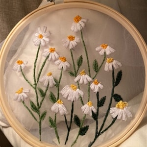 May include: White embroidered daisies with yellow centers on a sheer white fabric stretched over a wooden embroidery hoop.