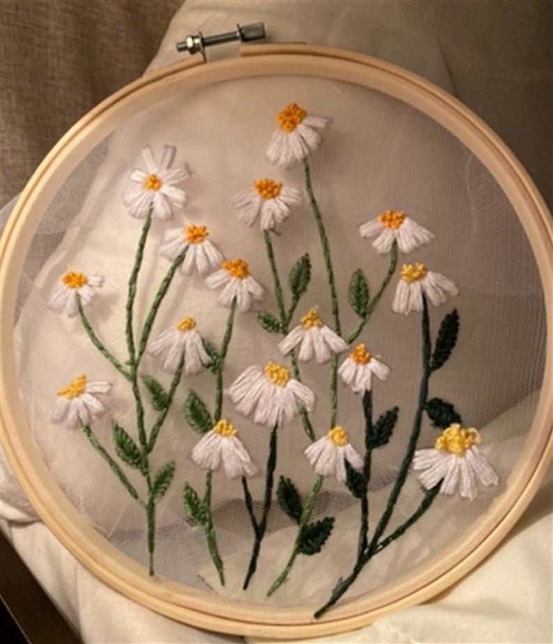 May include: White embroidered daisies with yellow centers on a sheer white fabric stretched over a wooden embroidery hoop.