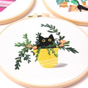 May include: Embroidered hoop art featuring a black cat peeking out of a yellow striped pot with green leaves and orange flowers.