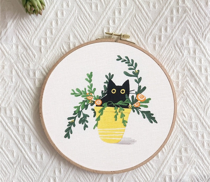 May include: Black cat embroidered in a yellow pot with green leaves and orange flowers. The embroidery is on a white circular canvas.