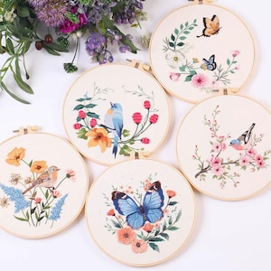 Könnte beinhalten: Eine Sammlung von fünf bestickten Reifen mit Naturmotiven. Jeder Reifen zeigt ein anderes Bild von Vögeln, Schmetterlingen und Blumen in leuchtenden Farben. Die Stickerei ist in einem Holzreifen angebracht.