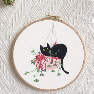 May include: Embroidered hoop art featuring a black cat peeking out of a pink pot with red flowers. The cat is hanging from a string.