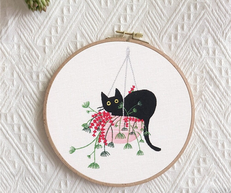 May include: Embroidered hoop art featuring a black cat peeking out of a pink pot with red flowers. The cat is hanging from a string.