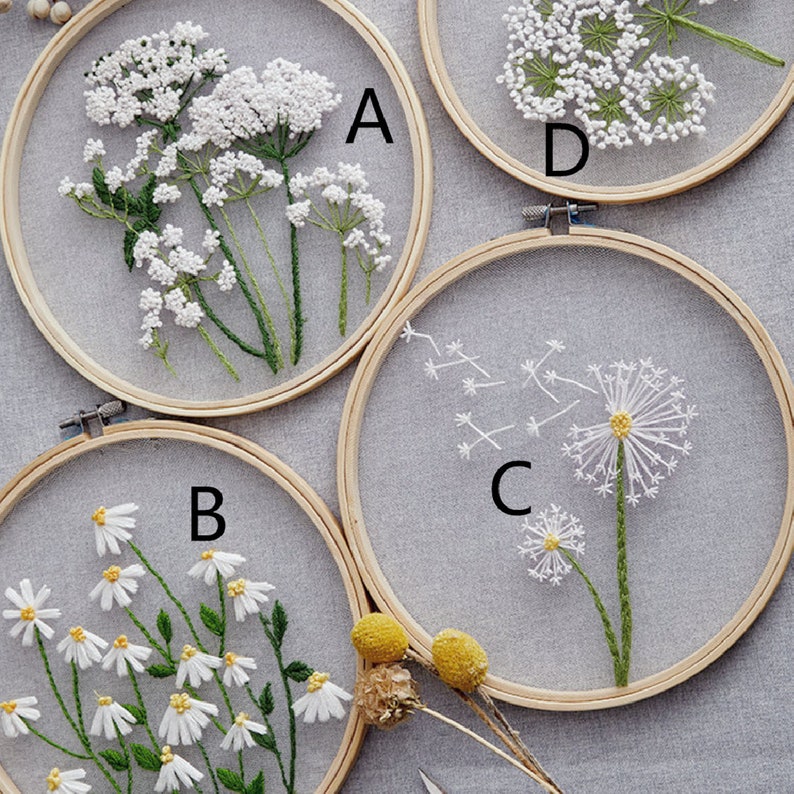 May include: Four embroidery hoops with different floral designs. The hoops are made of wood and have a white fabric stretched over them. The designs include a cluster of white flowers, a single dandelion, a cluster of white daisies, and a cluster of white flowers with a yellow center.