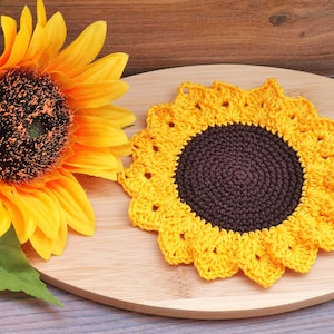 May include: A crocheted sunflower coaster with a brown center and yellow petals sits on a wooden surface next to a vibrant artificial sunflower. The coaster mimics the flower's design, perfect for home decor.