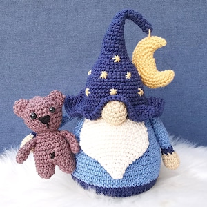 May include: A blue crocheted gnome wearing a hat with stars and a crescent moon. The gnome is holding a small brown crocheted teddy bear.