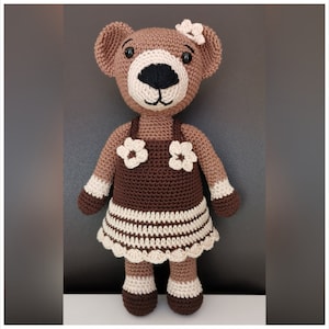 May include: A brown crocheted teddy bear wearing a brown and white dress with white flowers. The bear has black eyes and a black nose.