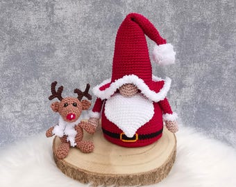 Puede incluir: Figuras navideñas de ganchillo: un gnomo y un reno. El gnomo es rojo con barba y borde de sombrero blancos. El reno es marrón con cuernos y bufanda blanca. Ambos están sobre una rodaja de madera.