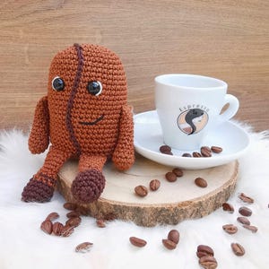Puede incluir: Un juguete de peluche de grano de café tejido a crochet se sienta en una rodaja de madera con granos de café esparcidos a su alrededor. Una taza de espresso blanca con la palabra "Espresso" impresa en ella se sienta en un platillo blanco.