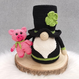 Może przedstawiać: Szydełkowy gnom na Dzień Świętego Patryka z czarnym cylindrem i zieloną koniczyną, obok różowej szydełkowej świnki z zielonym szalikiem. Gnom ma białą brodę i beżowy nos. Przedmioty znajdują się na drewnianym plastrze.