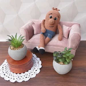 Könnte beinhalten: Eine gehäkelte Puppe mit braunem Körper, blauen Shorts und schwarzen Schuhen sitzt auf einem rosa Samtsofa. Zwei Topfpflanzen stehen auf dem Boden, eine mit grünen Sukkulenten und die andere mit einer grünen Pflanze mit langen, dünnen Blättern.