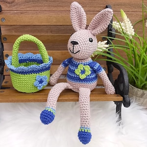 Könnte beinhalten: Ein gehäkeltes rosa Kaninchen, das einen blau-grünen gestreiften Pullover mit einer grünen Blume auf der Vorderseite trägt. Das Kaninchen sitzt auf einer Holzbank mit einem grün-blauen gehäkelten Korb.