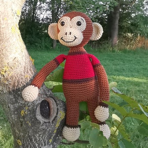 Puede incluir: Un juguete de mono de crochet con pelaje marrón, una camisa a rayas rojas y marrones y una cara amigable. El mono está sentado en un tronco de árbol.