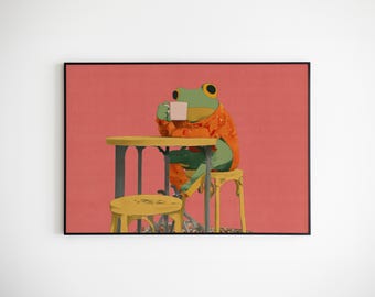 May include: A framed art print featuring a green frog wearing an orange sweater, seated at a yellow table and chair, holding a white mug. The background is a solid coral color. The frog appears to be enjoying a beverage.