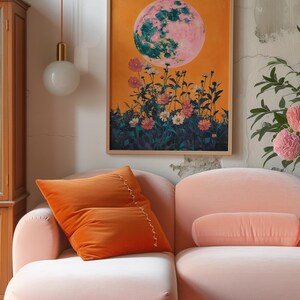 May include: A pink and green moon with a yellow background and flowers in the foreground. The moon is surrounded by a field of pink and white flowers. The image is framed and hanging on a wall above a pink sofa.