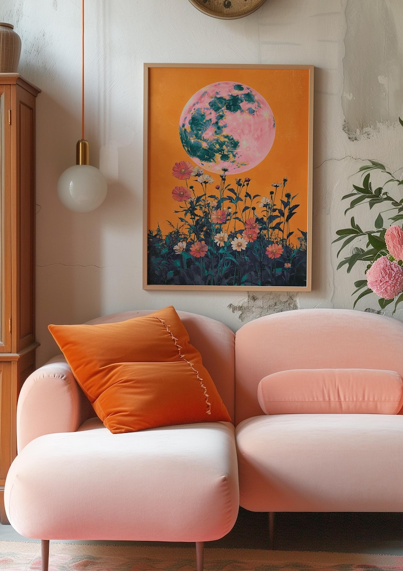 May include: A pink and green moon with a yellow background and flowers in the foreground. The moon is surrounded by a field of pink and white flowers. The image is framed and hanging on a wall above a pink sofa.