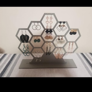 May include: A grey hexagonal earring stand with 12 compartments to display earrings. The stand is made of metal and has a modern design. The stand is filled with a variety of earrings in different colours and styles.
