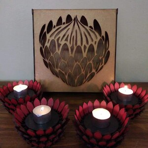 Puede incluir: Cuatro portavelas rojos con forma de flor, cada uno con una sola vela de té. Detrás de los portavelas hay una decoración de pared de madera con un diseño recortado de una flor de protea.