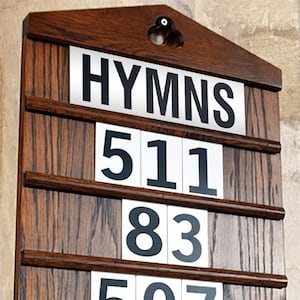 May include: A wooden hymn board displaying the word "HYMNS" at the top. Beneath are the numbers 511, 83, and 507 on white cards. The board is constructed from dark wood and features a decorative top.