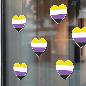 May include: Heart-shaped stickers displaying the non-binary pride flag. Each sticker has a yellow top, white middle, purple centre, and black base. The stickers are attached to a glass surface.