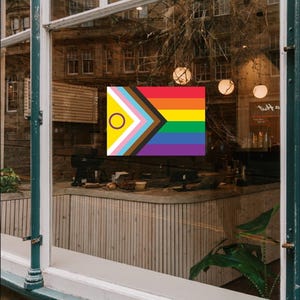 May include: A Progress Pride flag, incorporating the rainbow and additional colours, is displayed in a window. The flag's design includes colours representing transgender, Black, and brown communities, and a circle for intersex people. The flag is in front of a cafe.