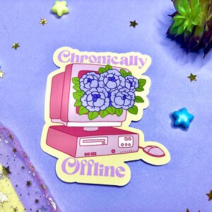 May include: A sticker featuring a pink retro computer with a bouquet of blue flowers on the screen. The words "Chronically Offline" are written in a light purple font. The sticker is on a purple background with star-shaped and star-shaped decorations.