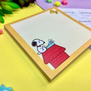 May include: A square sticky note pad with a light cream surface and a tan border. The bottom center features a graphic of the cartoon character Snoopy sitting on a red doghouse while typing on a small blue typewriter. The top edge has a small yellow icon.