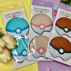 May include: Four round car coasters with a white background and a colorful design of a Pokeball. The coasters are packaged in clear plastic bags with a white header that reads "Car Coasters Handmade with love".