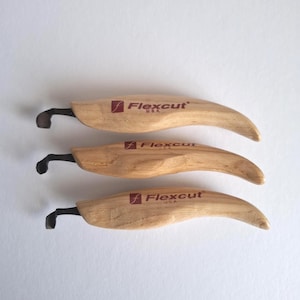 May include: Three wood carving tools by Flexcut USA, featuring curved blades. The handles are light brown wood, and the blades are black. The Flexcut logo is printed in red on the handle.
