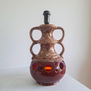 May include: A ceramic table lamp base featuring a brown and cream mottled glaze with two large handles on the sides. The lower section has a smooth dark brown finish with multiple circular cutouts that reveal a bright orange interior glow.