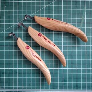 May include: Three wood carving tools with curved blades and wooden handles. Each tool has the "Flexcut USA" logo printed on the handle in red. The tools are arranged on a green cutting mat.