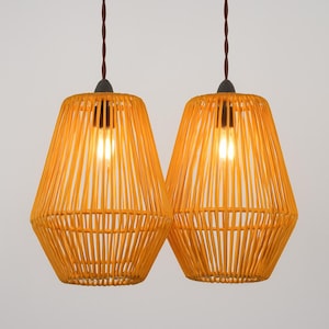 May include: Two matching pendant lights featuring bright orange, cage-style shades constructed from vertical rods. Each lamp is suspended by a dark brown twisted cord and contains a single glowing light bulb visible through the open structure.