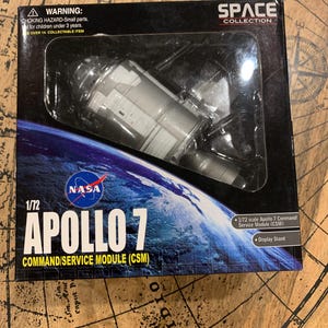 May include: A 1/72 scale model of the Apollo 7 Command/Service Module (CSM) in a black box with a white NASA logo and the text "Apollo 7" in yellow. The box also includes the text "Space Collection" and a warning about choking hazards.