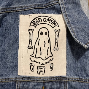 May include: A white fabric patch with black ink depicting a ghost with two bones and teeth, and the text "BAD OMEN" in a banner above the ghost.