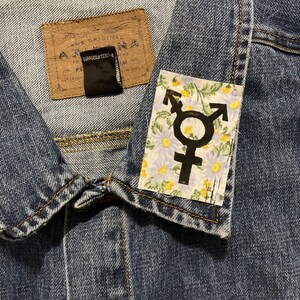 Op de afbeelding: Een blauwe denimjas met een patch op de kraag. De patch heeft een bloemenpatroon met een zwart transgender symbool in het midden.