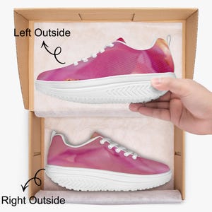 May include: A pair of pink and white athletic shoes with a textured design, white laces, and a thick white sole. The shoes are in a cardboard box, with one shoe held by a hand. The shoes are designed for exercise and fitness.