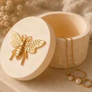 May include: A cream-coloured round jewellery box with a gold bee emblem on the lid. The box is open, revealing a gold necklace with pearls and a geometric pendant. The box has a honeycomb pattern.