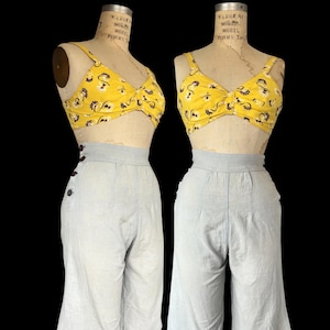 May include: A two-piece outfit consisting of a yellow crop top with a dark floral pattern and a twisted front detail, paired with light blue high-waisted culottes featuring a side button closure and visible stitching along the front seams.