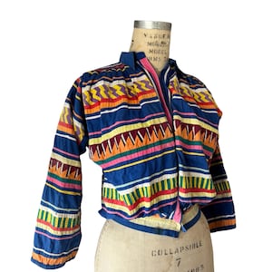 May include: A long-sleeved, cropped jacket featuring a vibrant, multi-colored geometric pattern on a deep blue background. The design includes horizontal stripes of yellow, orange, pink, and white zig-zags and solid bands, with a collared neckline.