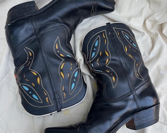 Könnte beinhalten: Ein Paar schwarze Leder-Cowboystiefel. Die Stiefel haben dekorative Nähte in Gelb, Blau und Rot. Die Stiefel haben eine spitze Zehenpartie und einen niedrigen Absatz. Die Stiefel werden auf einem cremefarbenen Stoff präsentiert.