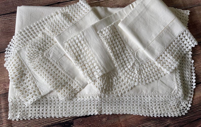 K&ouml;nnte beinhalten: Ein wei&szlig;es Leinentischtuch und passende Servietten mit aufwendiger H&auml;kelspitze. Das Set liegt auf einer Holzoberfl&auml;che und zeigt die filigrane Handwerkskunst und das klassische Design.